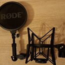 Černý popfilter s nápisem RØDE® a černý pavouk pro mikrofon. Oba doplňky jsou vyrobeny z kovu a plastu v matné černé barvě. Na spodní části držáku je…