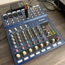 Modrý mixážní pult s nápisem YAMAHA MIXING CONSOLE MG10/2. Zařízení má řadu vstupních konektorů, regulační knoflíky v různých barvách a LED indikátor…
