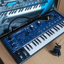 Modrý kompaktní syntetizátor s klávesnicí, na kterém je nápis MININOVA a logo novation. Za nástrojem stojí jeho originální krabice s nápisem Compact…