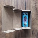 Modrý efekt pedál v kartonové krabici. Na přední straně jsou nápisy VOLUME, TONE, BOOST a AC STAGE ZIKZIC, doprovázené červeným grafickým motivem. Za…