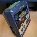 Černý trigger modul značky Roland s nápisem TRIGGER MODULE TM-2. Zařízení má malý displej, ovládací knoflík VOLUME a několik stlačovacích tlačítek na…