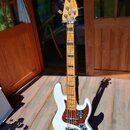 Bílá elektrická basová kytara s jasným dřevěným hmatníkem a červenoběle tečkovaným pickguardem. Na hlavě je nápis Fender JAZZ BASS. Tělo má stříbrný…
