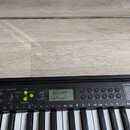 Černý digitální klavír s bílými a černými klávesami a kovovým notovým stojanem. Na ovládacím panelu je displej s nápisem 00 Piano 1 a 120 MELODY. Pan…