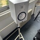 Bílý studiový monitor v hranatém korpusu umístěný na bílém trojnožném stojanu. Na přední straně jsou dva reproduktory a ve spodní části malý oválný n…