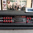 Černý basový zesilovač v textilním krytu s nápisem CARVIN R600 a RED LINE STEREO BASS AMP. Přední panel obsahuje řadu červených otočných ovladačů, po…