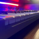 Detailní pohled na klávesy elektronického nástroje s bílými a černými klávesami. Nástroj je umístěn na tmavém dřevěném povrchu a osvětlen modrým a če…