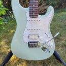 Světle zelená elektrická kytara typu Stratocaster s bílým perleťovým pickguardem a hnedým hmatníkem. Na pickguardu je podstranovými snímači nápis "Sq…