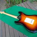Elektrická kytara s tvarem typu Stratocaster v barevném přechodu od žluté přes oranžovou k červené a černé. Zadní strana korpusu obsahuje bílý plasto…