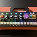 Černý elektronický kontrolér s řadou bílých podsvícených tlačítek očíslovaných 1 až 14. Zařízení obsahuje několik otočných potenciometrů, červené tla…