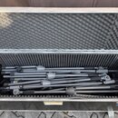 Otevřený černý transportní kufr s vnitřním polstrováním z šedé pěny. Uvnitř se nachází několik rozložených a složených šedých kovových stativů s čern…