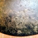 Detailní pohled na povrch kovového činele s výraznou patinou a oxidací. Na povrchu je vyryto logo znázorňující stylizovanou tvář a nápis BM-HR12. Pov…