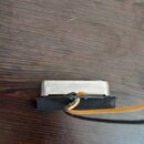 Fotografie zachycuje elektrický snímač s kovovým krytem stříbrné barvy. Snímač je připevněn k černému plastovému nebo gumovému podkladu, ze kterého v…