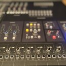 Detailní pohled na ovládací panel černého mixážního pultu s řadou posuvníků, knoflíků a tlačítek. Na panelu jsou vidět nápisy MAIN VOLUME, SOLO, CUT,…
