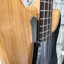 Detailní záběr na tělo a hmatník elektrické basové kytary z přírodního světlého dřeva. Nástroj má černý pickguard a hmatník z tmavého dřeva s kovovým…