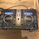 Černý DJ kontrolér s dvěma jog-koly a centrálním mixážním sekcí. Na horní hraně je bílý nápis DENON DJ a pod ním model MC4000. Zařízení obsahuje modř…
