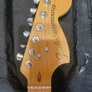 Fotografie zachycuje hlavu dřevěné elektrokytary v přirozené světle, umístěnou v černém pouzdře. Na dřevěném povrchu je nápis Fender STRATOCASTER a v…