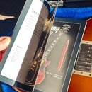 Fotografie zachycuje otevřený produktový manuál položený na modrém plyšovém vnitřku kufru. V pravé stránce je vidět obrázek červené elektrické kytary…