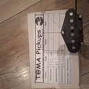 Černý elektrický snímač typu humbucker s viditelnými póly. Vedle snímače je položen papír s nápisem TUMA Pickups a tabulkou, kde je v řádku Bridge uv…