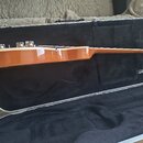 Kytara s naturalním dřevěným povrchem v černém tvrdém kufru. Na vnitřní straně kufru je vidět bílý nápis Gibson. Nástroj je zobrazen z profilu, jsou…