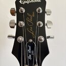 Černý hlavník elektrické kytary se stříbrnými ladičkami. V horní části je bílý nápis Epiphone a uprostřed zlatý nápis Les Paul MODEL. Na černém trsac…