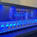 Kytarový zesilovač s černým rámem a průhledným předním panelem, vnitřní prostor je modře podsvícen. Uvnitř jsou vidět elektronické komponenty a lampy…