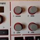 Detailní pohled na bílý MIDI kontrolér s nápisem MPKmini. Na panelu jsou šesti šedé otočné knoflíky s označením 1 DIVISION, 2 SWING, 3 MODE, 5 LATCH,…
