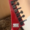 Záběr na červenou hlavu elektrické kytary s šesti černými ladicími kolíky. Na každém kolíku je bílý nápis jackson. V dolní části hlavy je bílá nálepk…