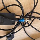 Černý napájecí adaptér s připojenými kabely. Na spodní straně adaptéru je nápis „LUTIONS“, „INPUT: 100-240V ~ 50/60Hz“, „OUTPUT: DC 9V 1A“ a další te…
