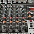 Stříbrošedý mixážní pult s řadou ovládacích prvků, včetně modrých a oranžových otočných knoflíků. V horní části je digitální displej s červenými čísl…