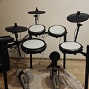 Elektronická bicí souprava s černým rámem a bílými padovými plochami. Na pedálech je viditelný nápis ALESIS. Soubor obsahuje čtyři pady, tři činely a…
