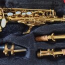 Zlatý saxofon v černém polstrovaném kufru. Vedle nástroje jsou umístěny tři příslušenství: černý mouthpiece s kovovým ligaturem a dva zlaté nástavce…