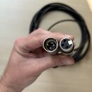 Ruka drží dva konce černého kabelu s konektory typu XLR. Na pravém konektoru je vidět nápis "MADE IN USA". Povrch kovových konektorů vykazuje mírné z…