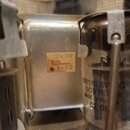 Fotografie zachycuje kovový transformátor stříbrné barvy. Na jeho boku je nalepený béžový štítek s nápisy 784.389, Drake Transformers a 1.90 s červen…