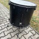 Černý buben s texturovaným povrchem a kovovými obručemi. Na horním obruči je viditelné logo značky Tama. Buben stojí na kovovém stojanu s černými gum…