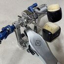 Detailní pohled na kovový mechanismus bubenového pedálu v stříbrné barvě s modrými anodizovanými prvky. Na kovové části je vyryto logo ve tvaru styli…