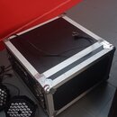 Černý transportní case s hliníkovými hranami a kovovými rohy. Na horní desce leží černý napájecí kabel s kolíkovým konektorem. Vedle case jsou vidět…