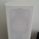 Bílý hranatý reproduktor s textilní maskou přes celý přední panel. V pravém dolním rohu je umístěno černé logo AQ. Zařízení stojí na tmavě hnědém pod…