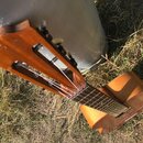 Dřevěná akustická kytara v hnědé barvě, opřená o šedý kovový předmět. Nástroj vykazuje známky opotřebení, jsou vidět škrábance a odřeniny na povrchu…