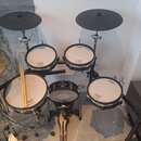 Elektronická bicí souprava s černými rámy a bílými membránami. Na membránách jsou vidět nápisy Roland a V-Drums. Souprava obsahuje čtyři pady, dva če…