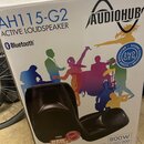 Fotografie znázorňuje bílou papírovou krabici s produktem Active Loudspeaker. Na krabici jsou nápisy AH115-G2, AUDIOHUB a logo Bluetooth. V pravé čás…