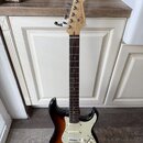 Elektrická kytara typu Stratocaster s barevným přechodem korálovo-černé (sunburst) a bílým pickguardem. Na hlavě hmatníku je vidět nápis Fender STRAT…