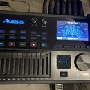 Černý elektronický modul s nápisem ALESIS a označením STRIKE PERFORMANCE DRUM MODULE. Zařízení obsahuje displej s modrým rozhraním, několik ovládacíc…