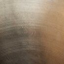 Detailní záběr na povrch kovového činele zlatavé barvy s výrazným koncentrickým rýhováním. Uprostřed je vidět vyryté logo s nápisem "Zildjian". Povrc…