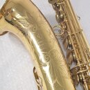 Detail zlatého saxofonu s ornamentálním rytím na čase. Uprostřed rytiny je nápis „MADE IN JAPAN NAGI KAW A TOKYO MODEL 54“. Povrch nástroje je lesklý…