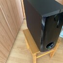 Černý, lesklý subwoofer s hranatým tvarem a oválným otvorem pro basreflex v dolní části. Na spodním okrahu je viditelné malé stříbrné logo LG. Zaříze…