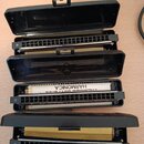 Tři otevřené černé plastové pouzdra s harmonikami. Na prostřední je vidět nápis HARMONICA a pod ním obráceně TREMOLO DIATONIC. Na pouzdrech jsou vidě…