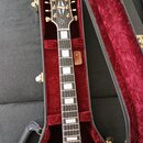 Elektrická kytara v červené barvě umístěná v otevřeném kufru s červeným plyšovým vnitřkem. Na hlavě hmatníku je zlatý nápis Gibson a pod ním bílý náp…