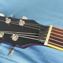 Detail hlavy elektrické kytary v tmavě fialové barvě s nápisem Vintage v žlutém kurzívním písmu. Na hlavě jsou šest kovových ladiček a bílá matice. H…