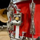 Detail červeného bubnu s chromovaným kováním. Na korpusu je připevněn držák s nápisem YAMAHA, který drží bílý textilní popruh. Povrch nástroje je glo…