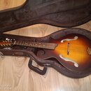 Akustická archtop kytara s barevným přechodem od žluté k tmavě hnědé, umístěná v hnědém tvrdém kufru s polstrováním. Na hlavě nástroje je viditelné z…